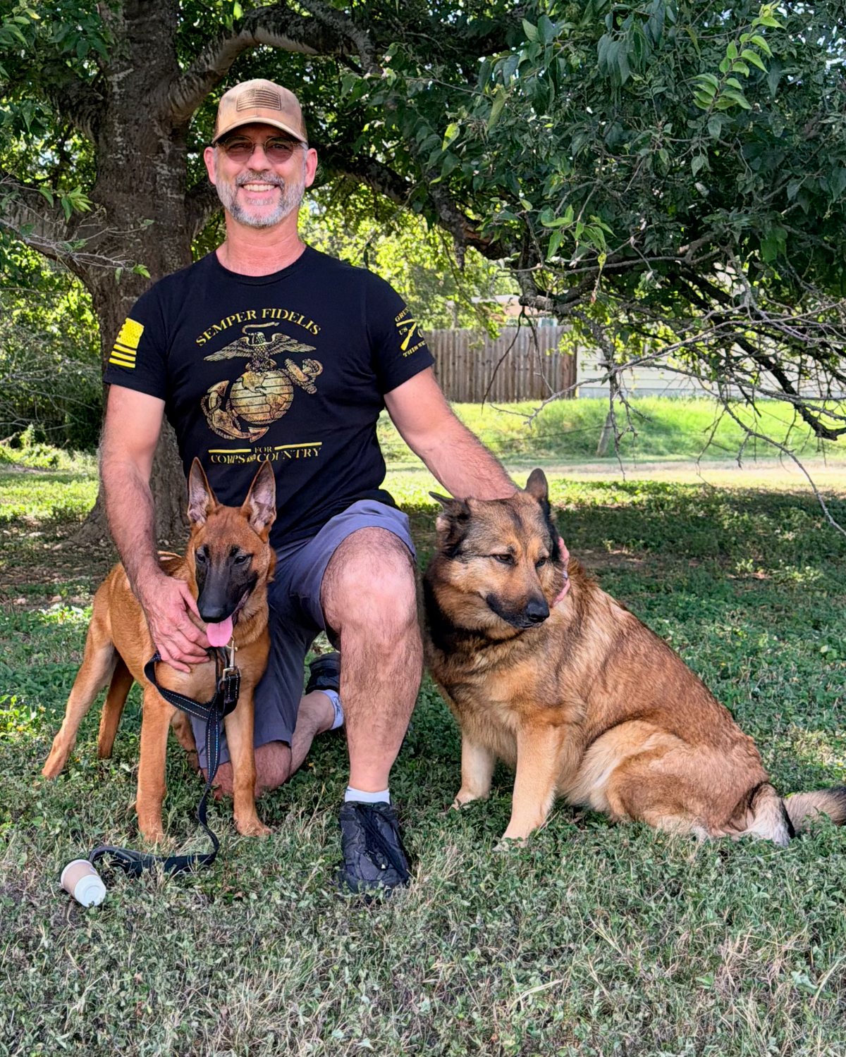 Rusty Nokes kneeling under a pecan tree with his two dogs — River, a German Shepherd, and Lady, a young Belgian Malinois — both sitting calmly beside him