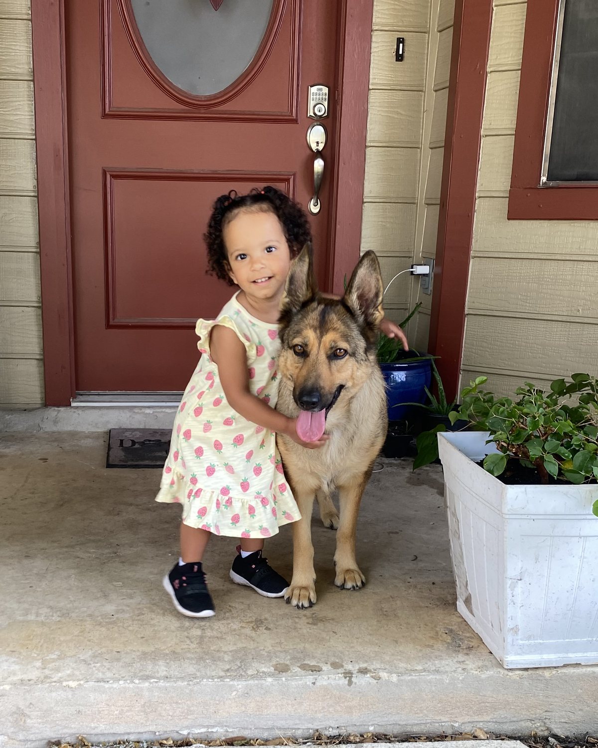 A toddler in a strawberry-print dress hugging River, a German Shepherd, on a front porch