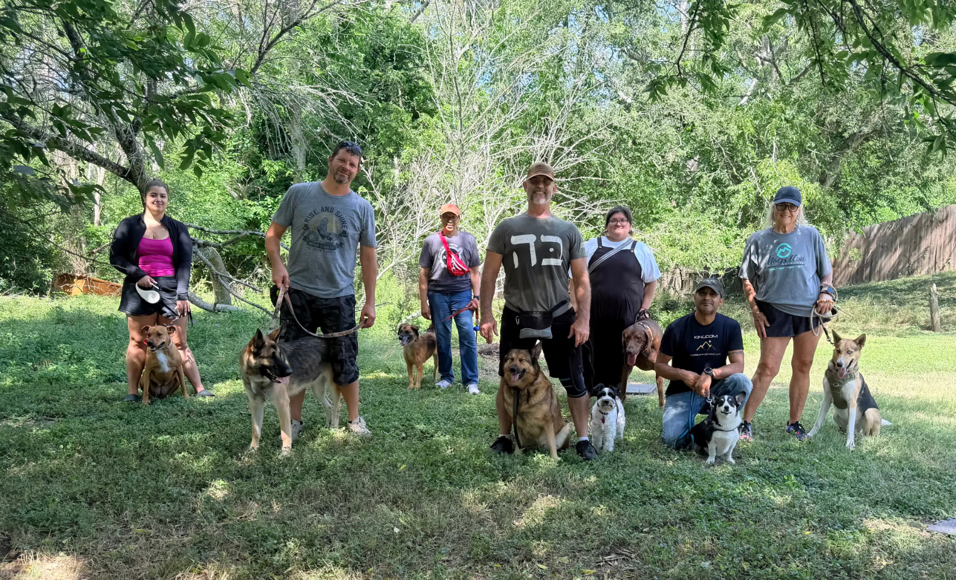 A group obedience boot camp session: handlers spaced across a clearing holding their dogs in place-and-stay positions.